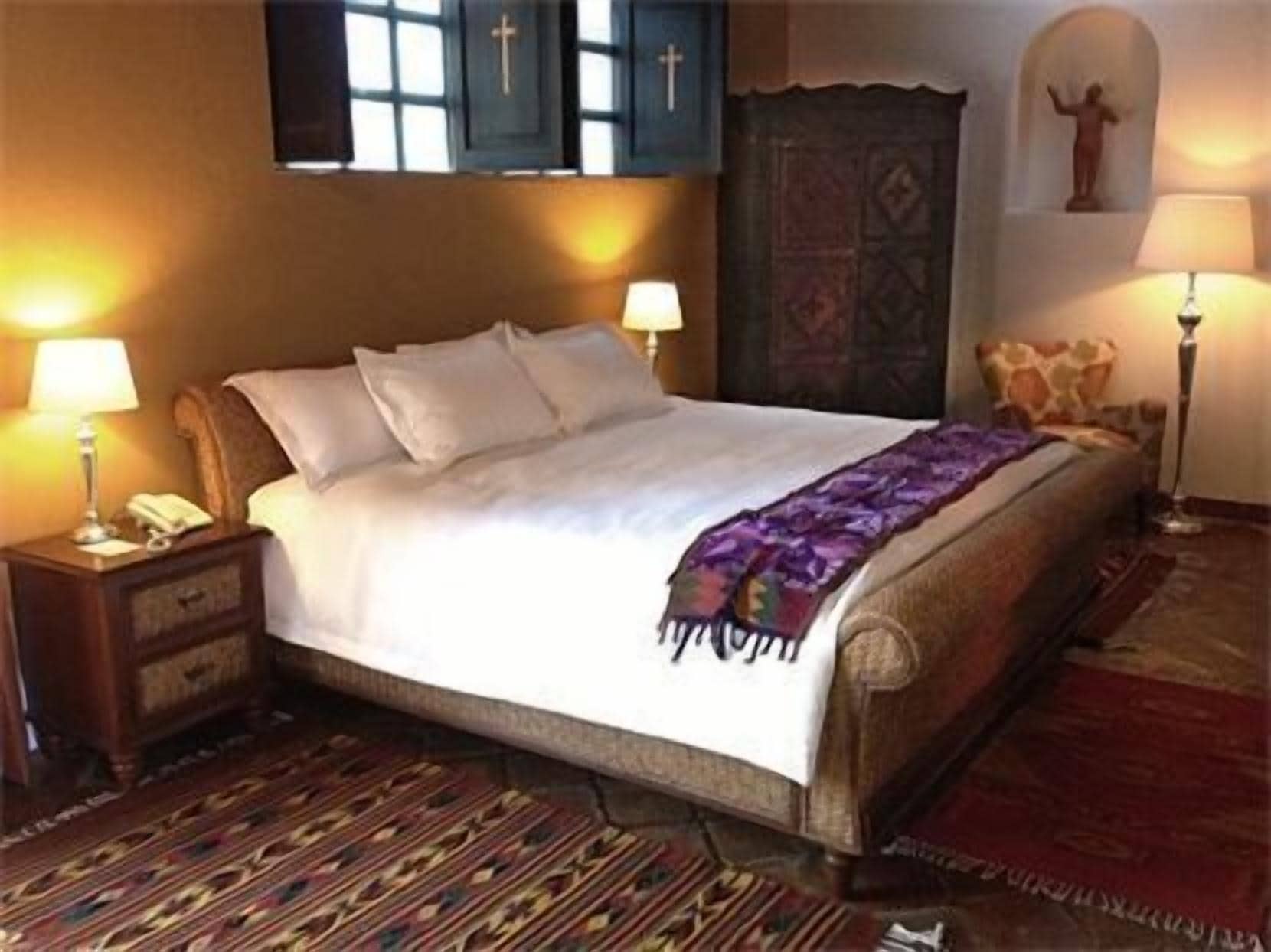 A neatly made bed with a white comforter and pillows, a wooden nightstand with a lamp, a patterned rug, and a religious statue on the wall.