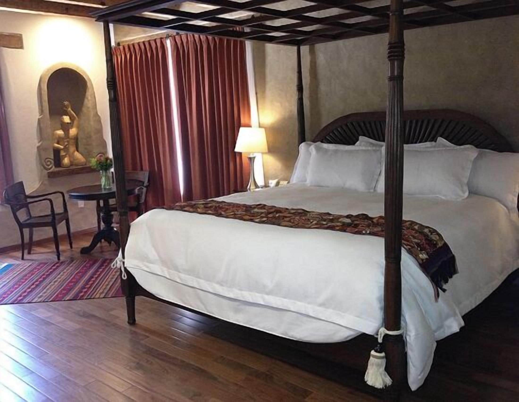 A four-poster bed with white linens, a wooden headboard, and a decorative throw. There is a small table with a lamp and a chair to the left.