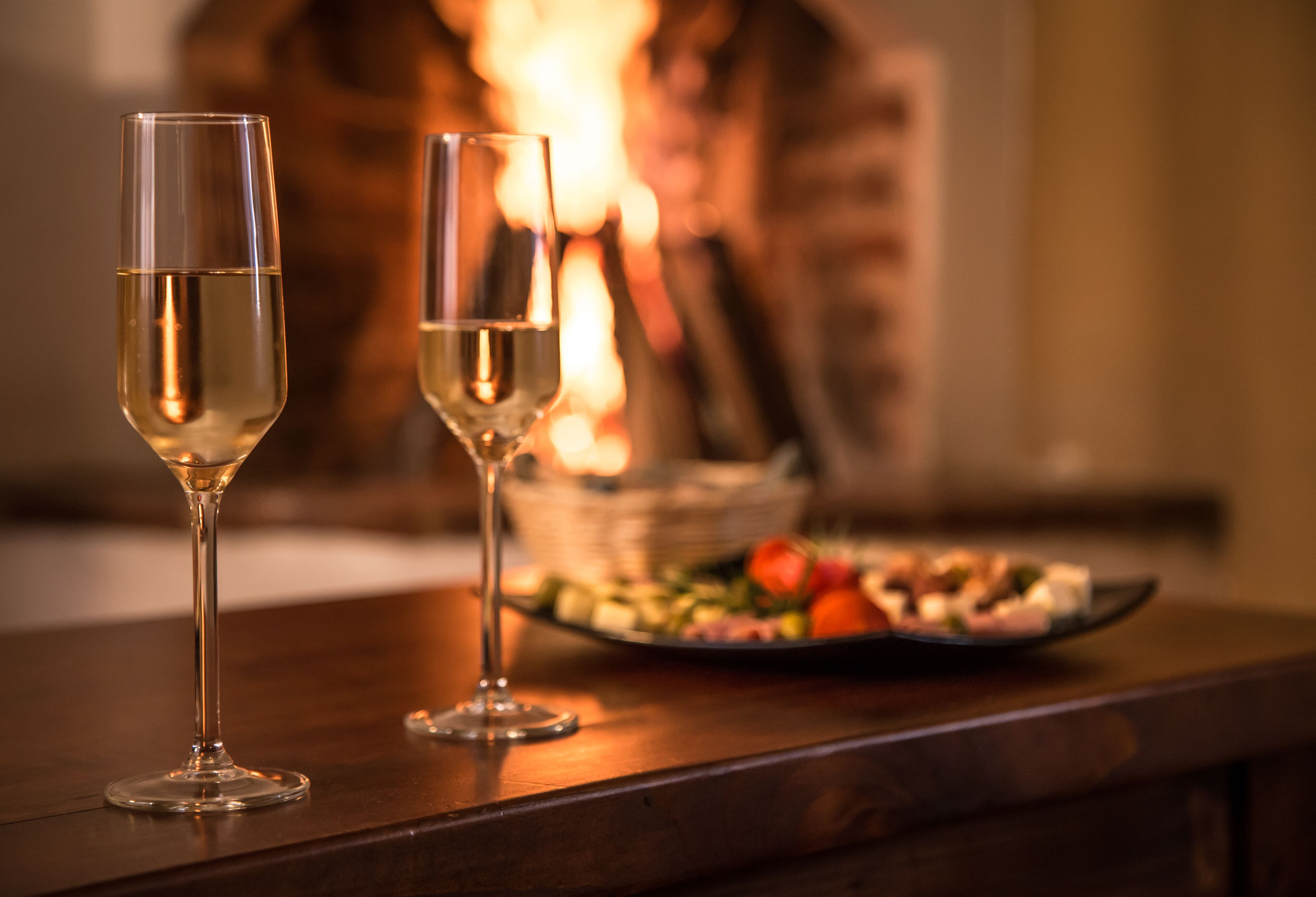 Two glasses of white wine on a wooden table with a plate of food and a fireplace in the background.