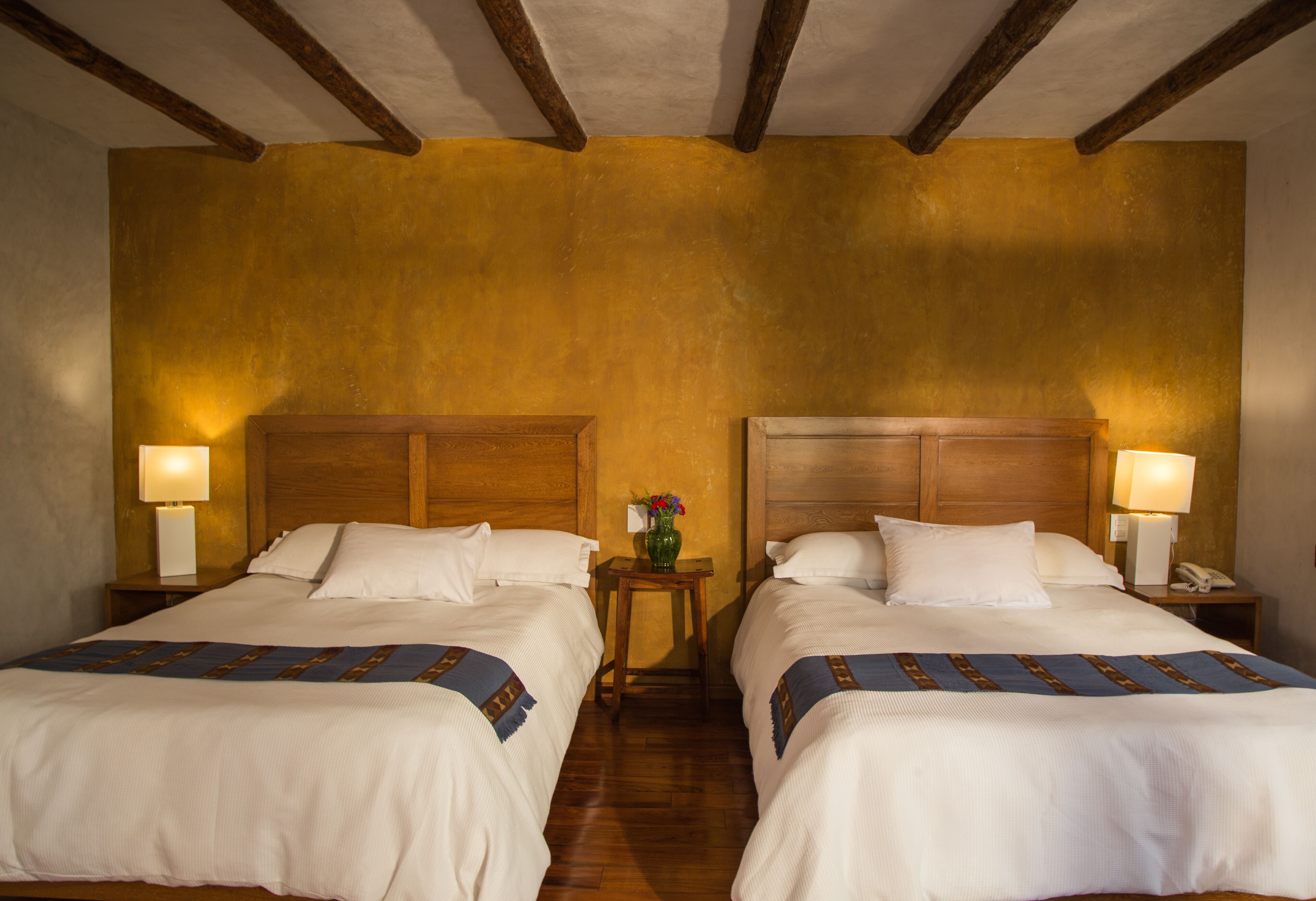 A hotel room with two beds, wooden headboards, bedside lamps, a small table with a vase of flowers, and a wooden floor.