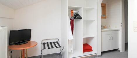 A compact room with a television, a small table, a metal drying rack, a wardrobe with a red item, and a bathroom with a sink and cabinets.