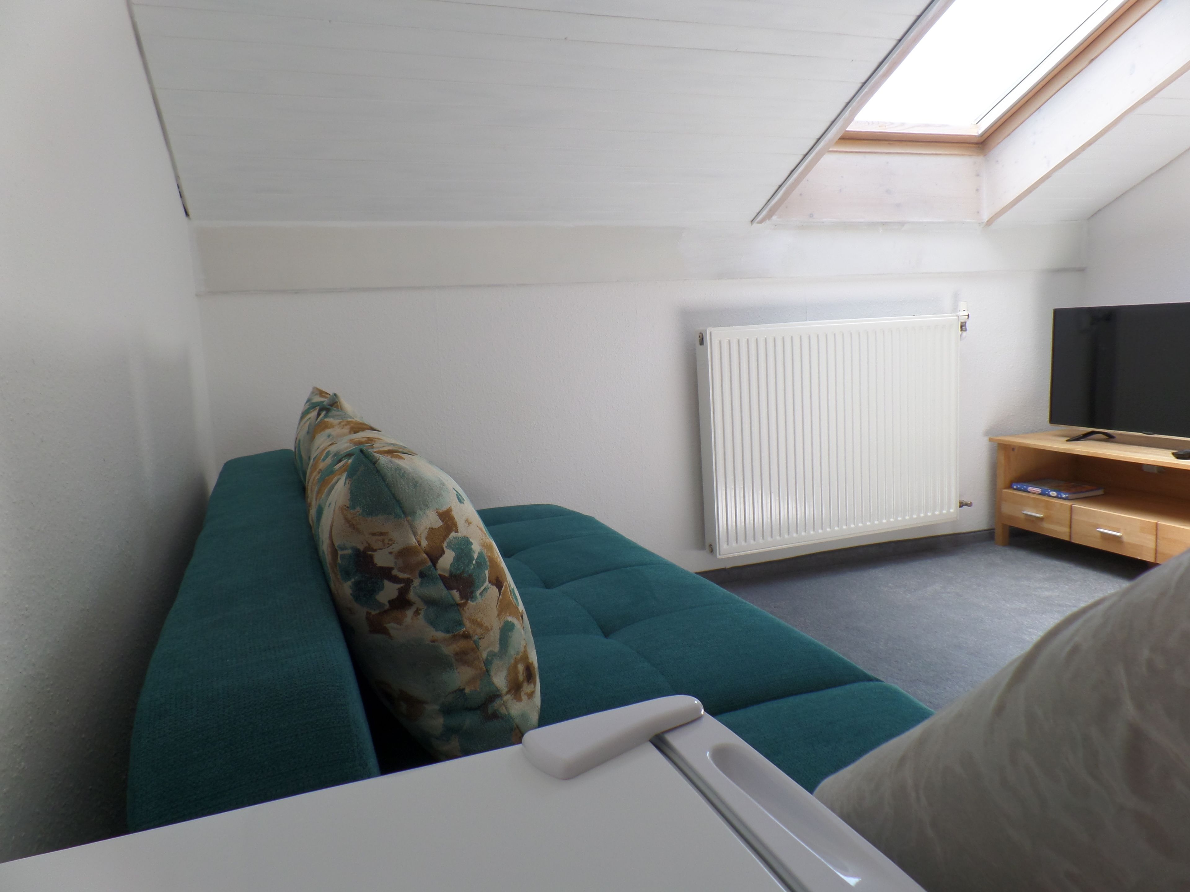 Ein kleiner, gemütlicher Raum mit einem türkisfarbenen Sofa, einem Kissen, einem weißen Tisch, einem Holz-TV-Schrank und einem Dachfenster.