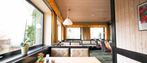 A restaurant with a wooden ceiling and large windows. There are tables and chairs, and a potted plant on the windowsill.