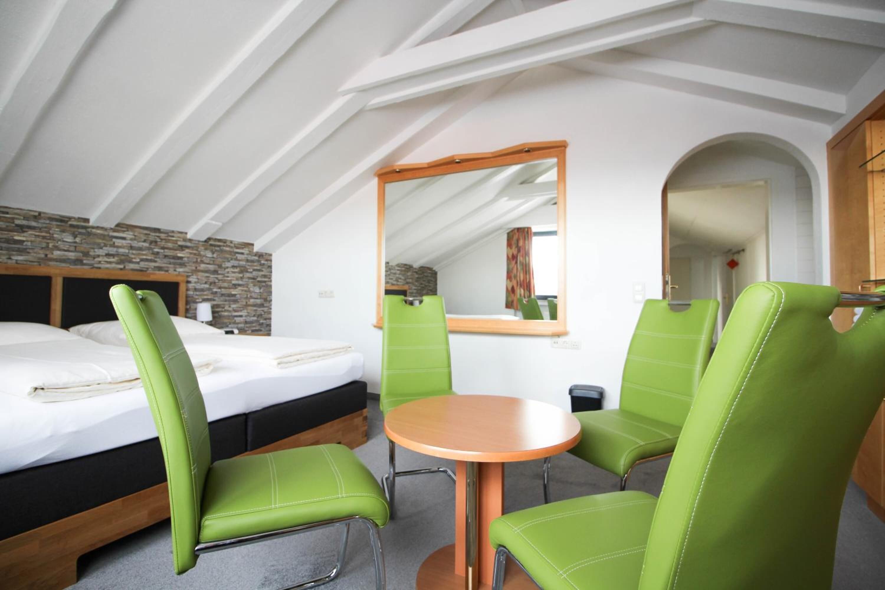 A room with a bed, green upholstered chairs, a round wooden table, and a mirror on the wall.