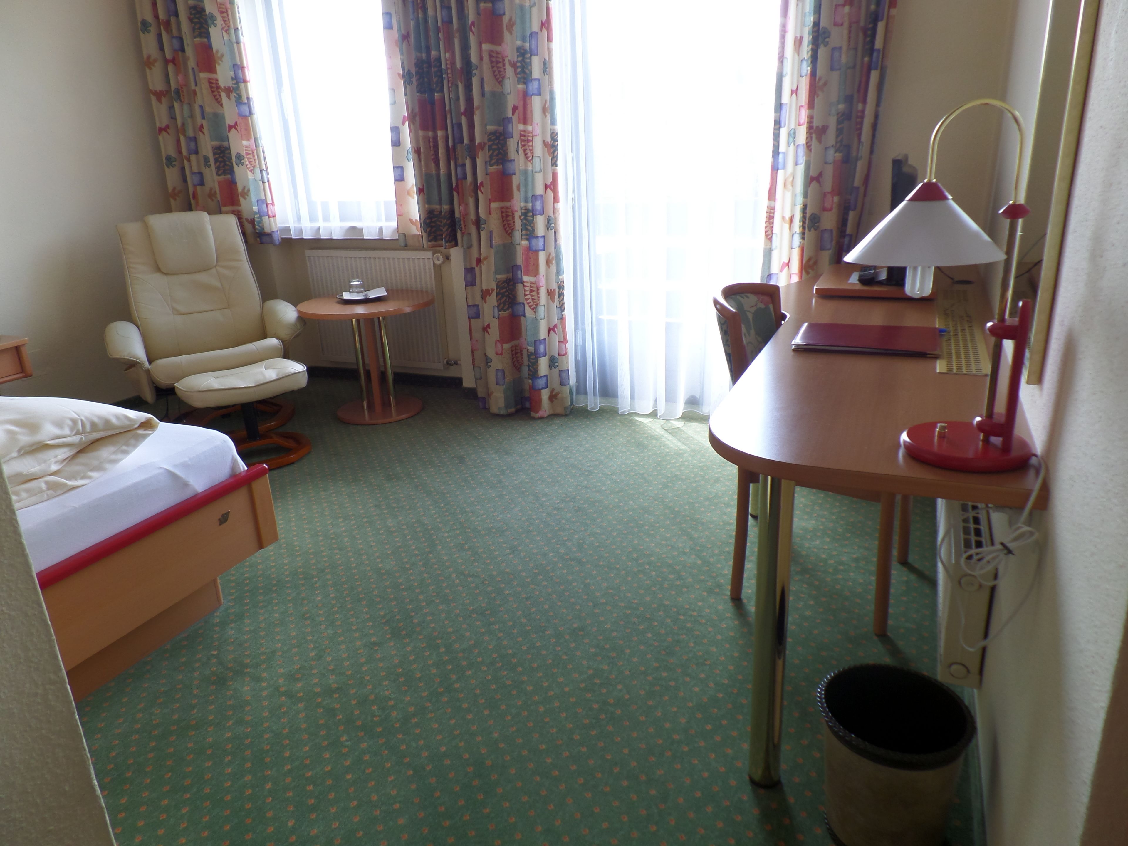 Hotelkamer met een bed, een stoel, een tafeltje, een bureau met een lamp en een radiator.