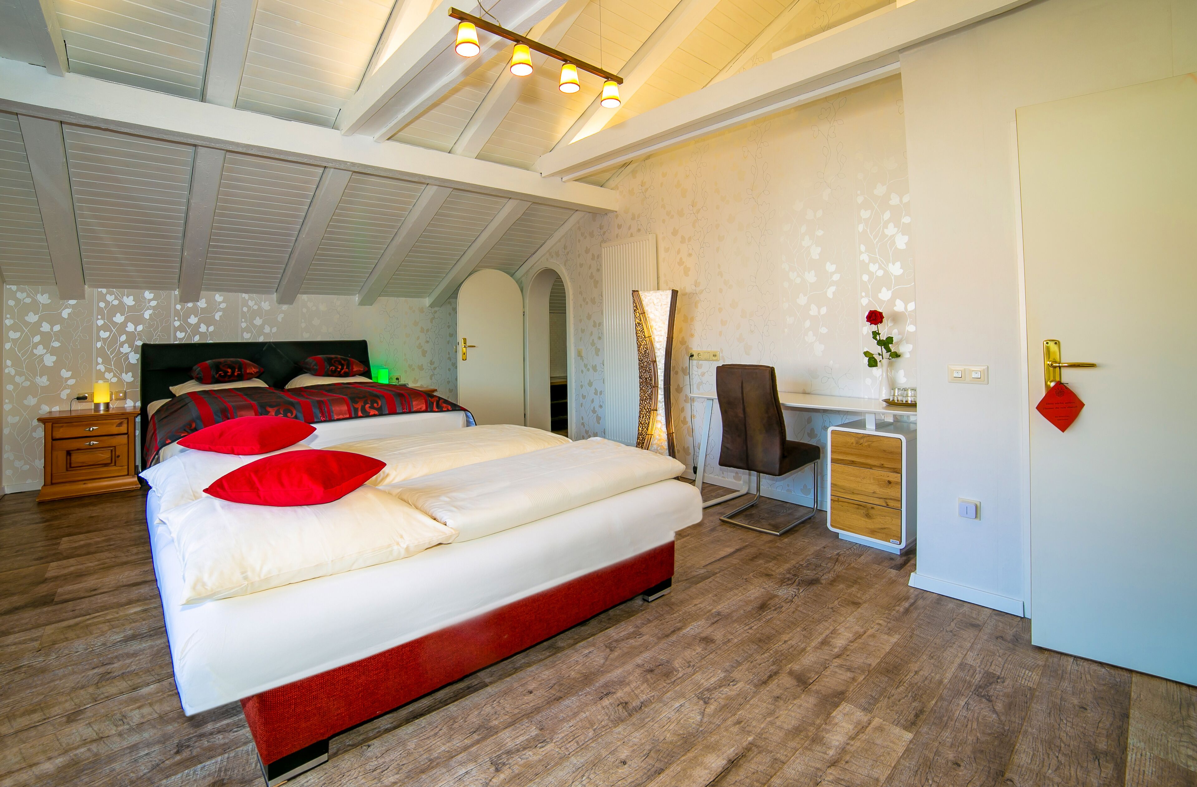 A bedroom with a large bed, a desk, and a chair. There is a wardrobe and a door with a red decoration.