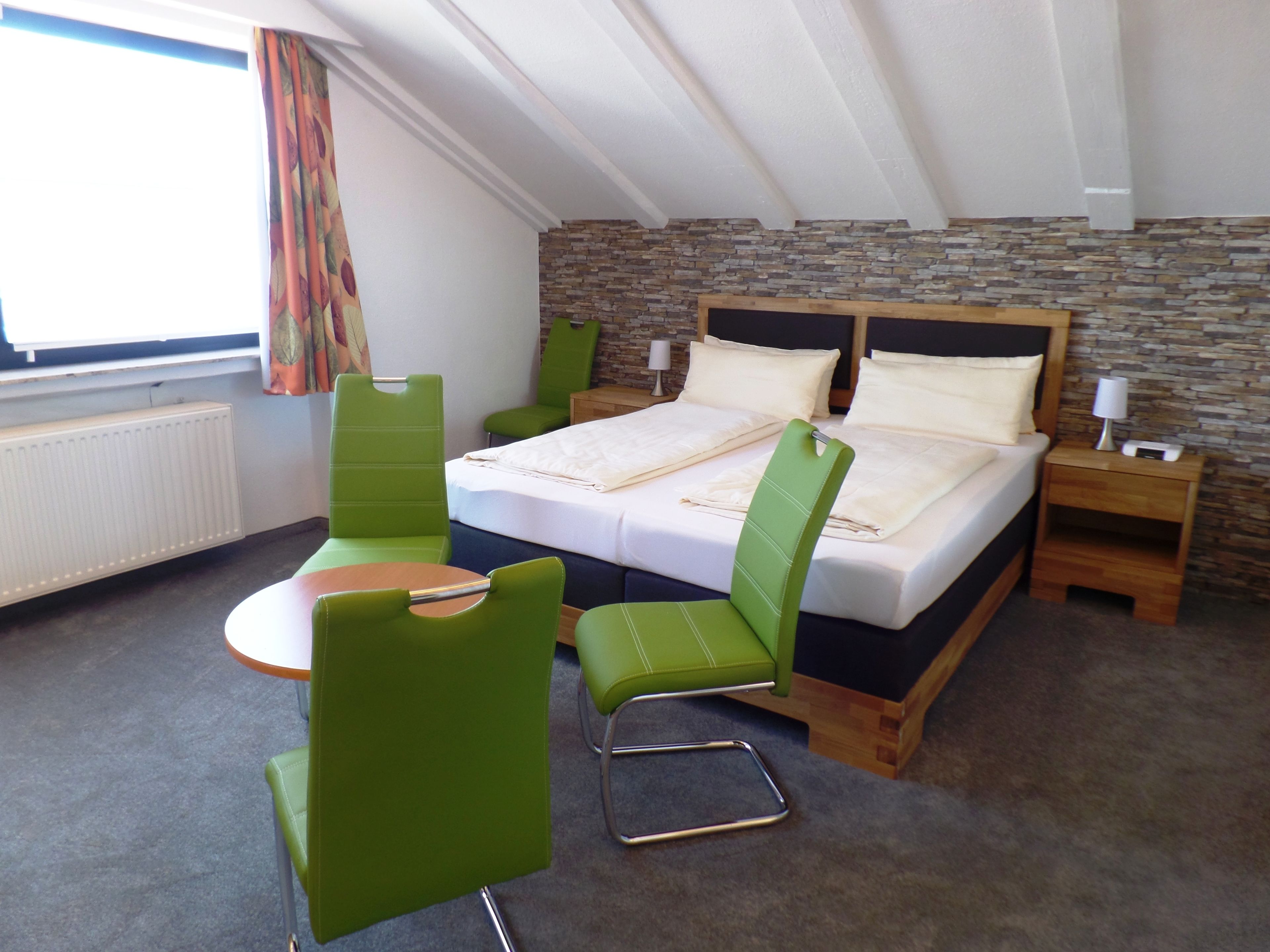 A hotel room with a bed, two green chairs, a small table, a nightstand with a lamp, and a stone wall.