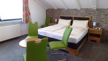 A hotel room with a bed, two green chairs, a small table, a nightstand with a lamp, and a stone wall.