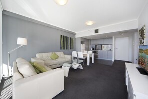 A modern living room with a sofa, coffee table, and dining area. There is a kitchen in the background with cabinets and appliances. - Astra Apartments - Paramatta (Parramatta)