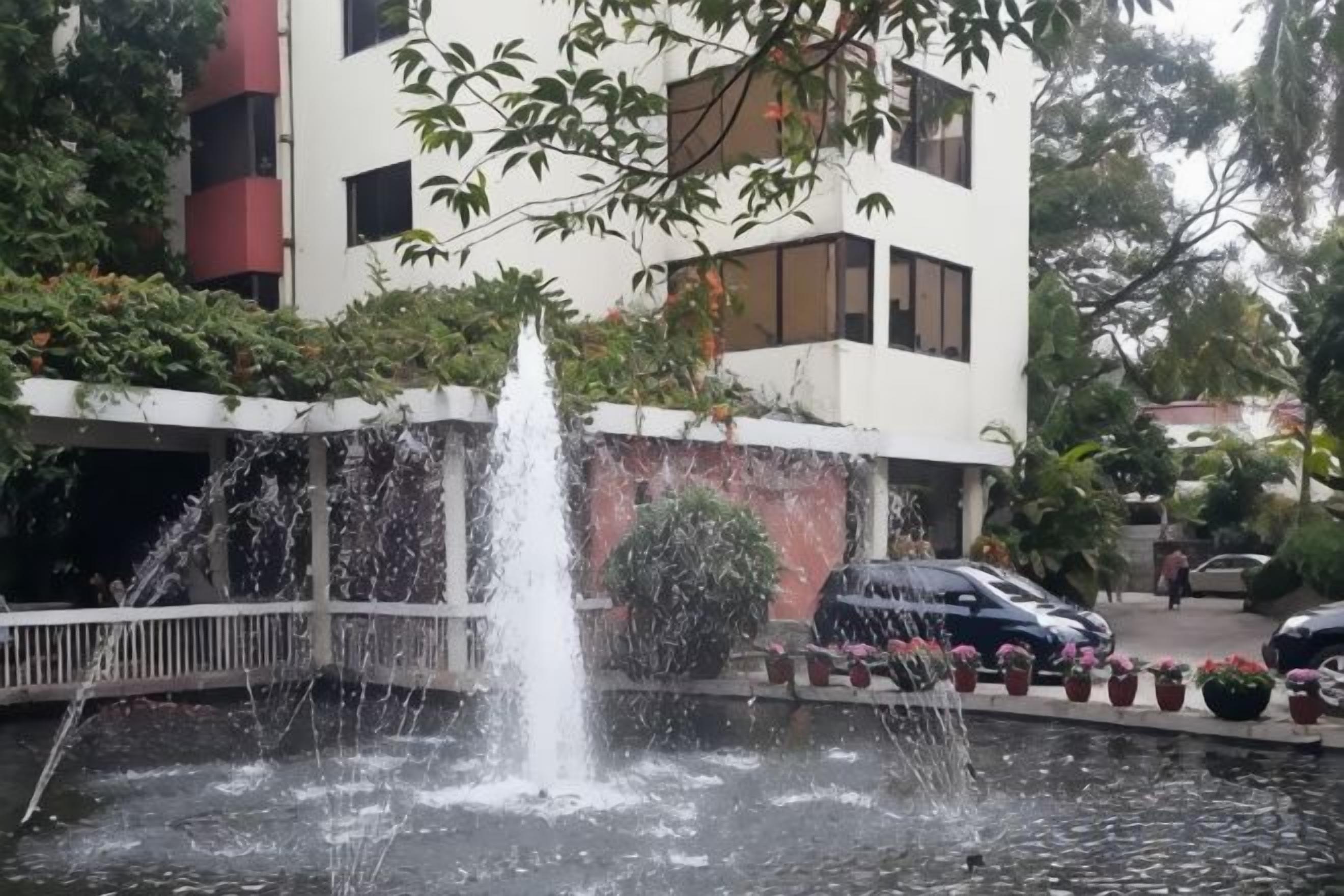Una fuente con chorros de agua, un estacionamiento con automóviles y un edificio con balcones.