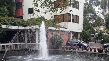 Una fuente con chorros de agua, un estacionamiento con automóviles y un edificio con balcones.