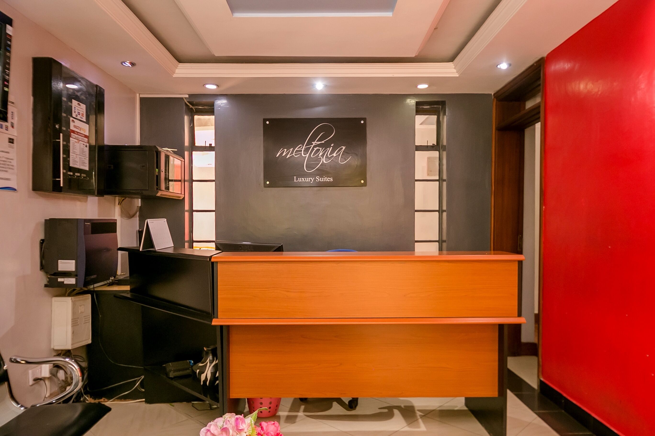 A reception desk with a sign reading "Melgia Luxury Suites," a black cabinet, a computer, and a red wall.