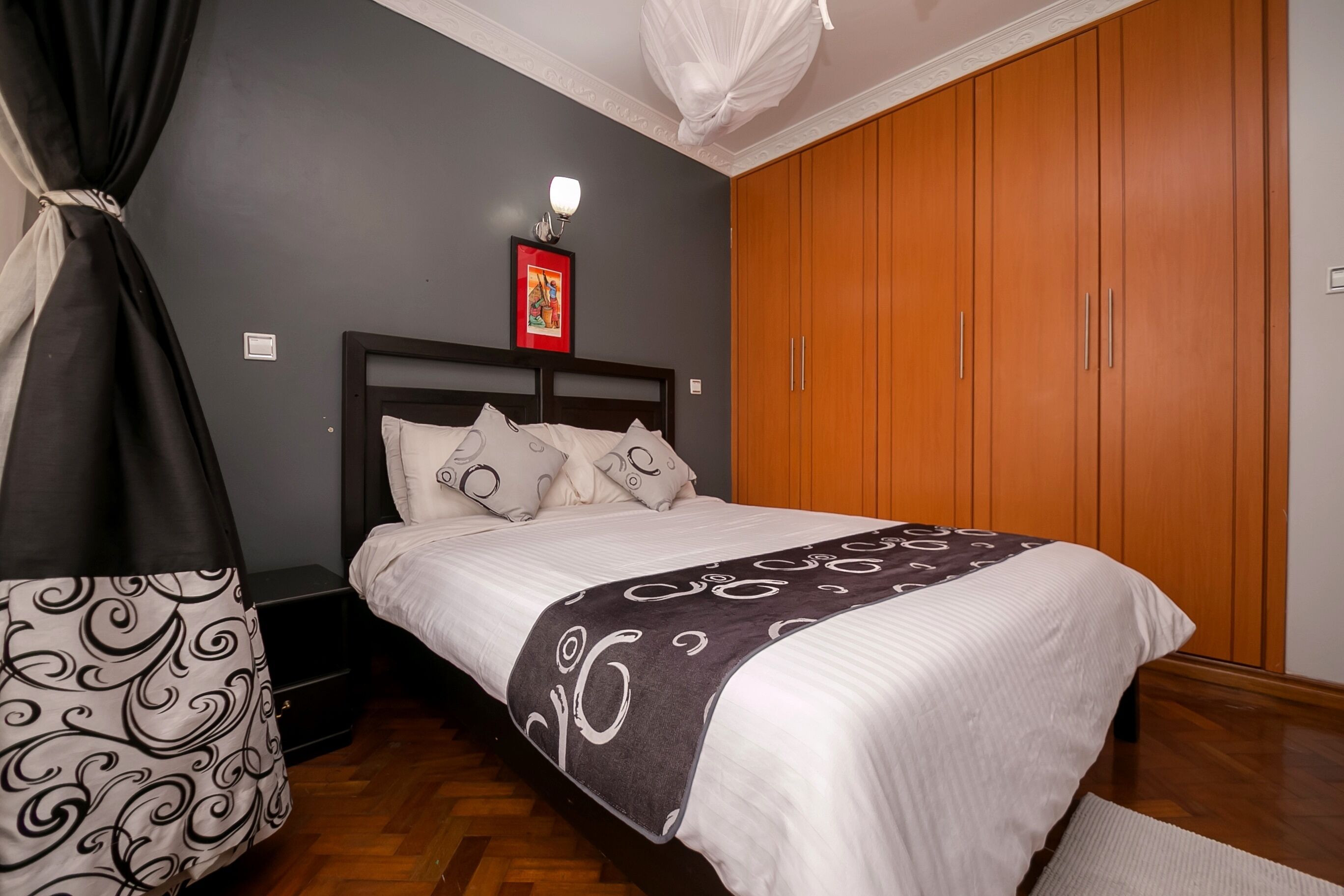 A neatly made bed with a white and gray patterned blanket, a black and white patterned bag, a wooden wardrobe, and a wall-mounted light fixture.