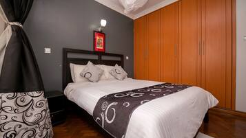 A neatly made bed with a white and gray patterned blanket, a black and white patterned bag, a wooden wardrobe, and a wall-mounted light fixture.