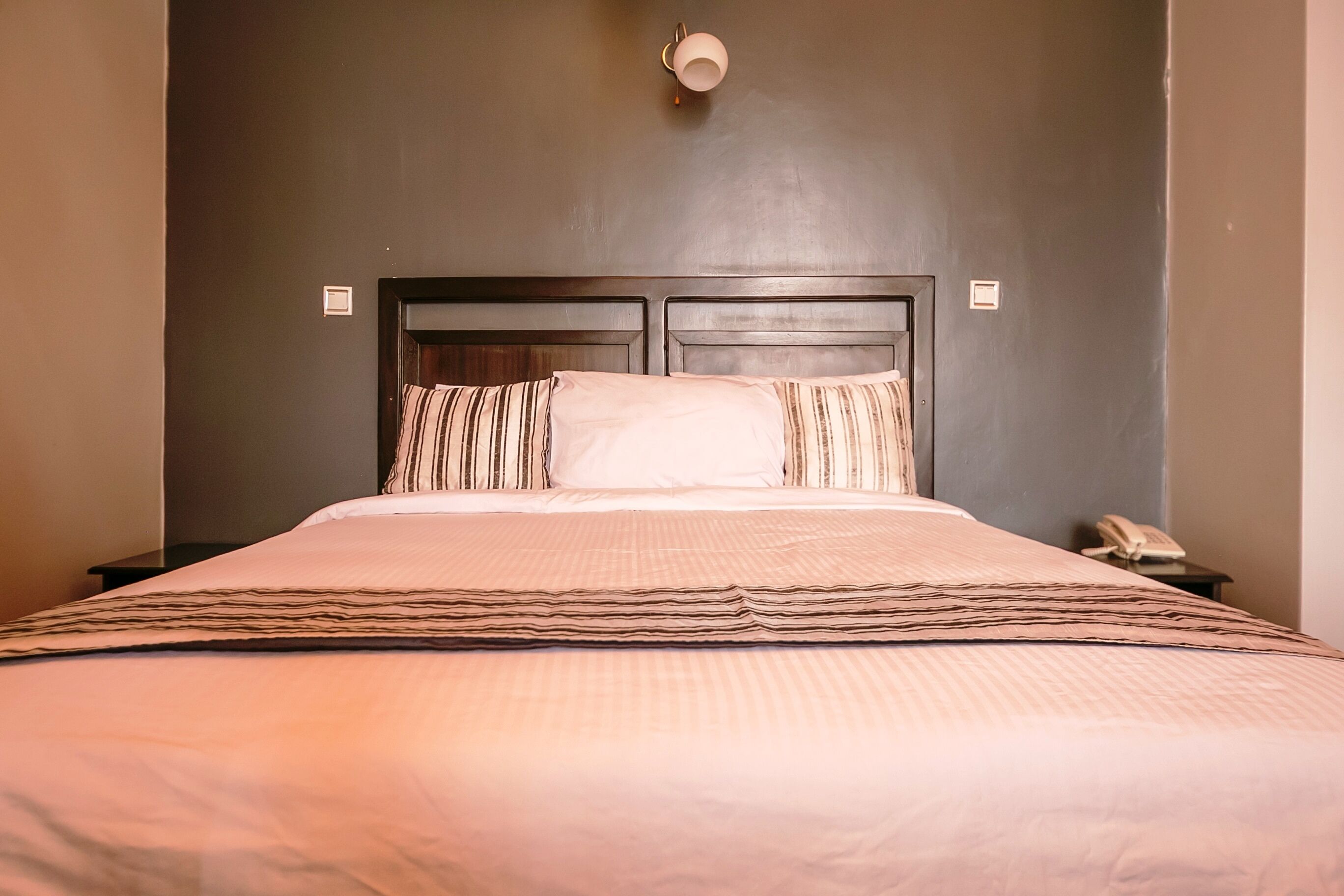 A neatly made bed with striped pillows and a headboard, a bedside phone, and a wall-mounted light fixture.