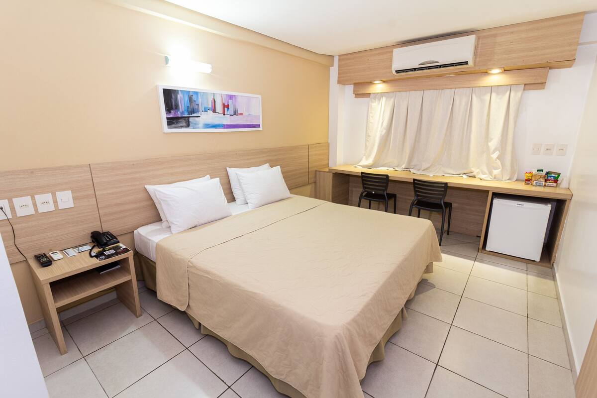 A hotel room with a bed, bedside table, desk, chair, and a small refrigerator.