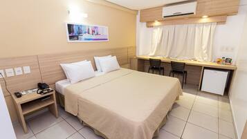 A hotel room with a bed, bedside table, desk, chair, and a small refrigerator.