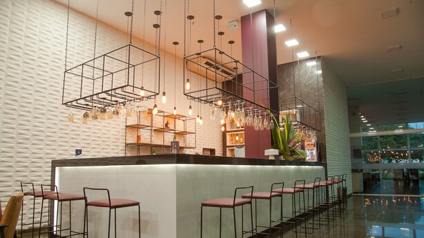 A modern bar area with a long counter, multiple stools, hanging light fixtures, and a patterned wall.