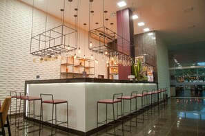 Zona de bar moderna amb una barra llarga, diverses solles, lluminàries penjants i una paret amb un disseny decoratiu.