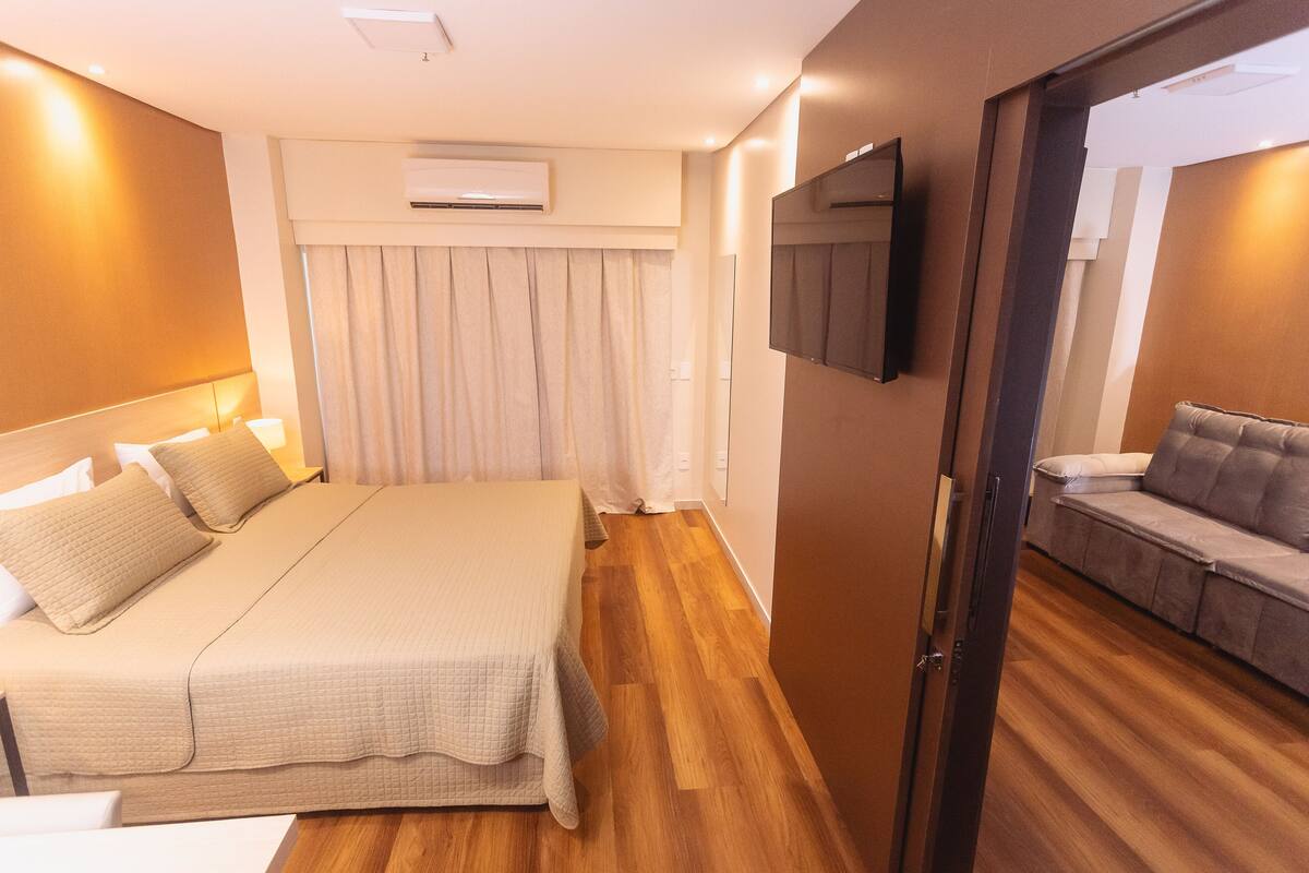 A modern hotel room with a large bed, a sofa, a flat-screen TV, and an air conditioning unit.