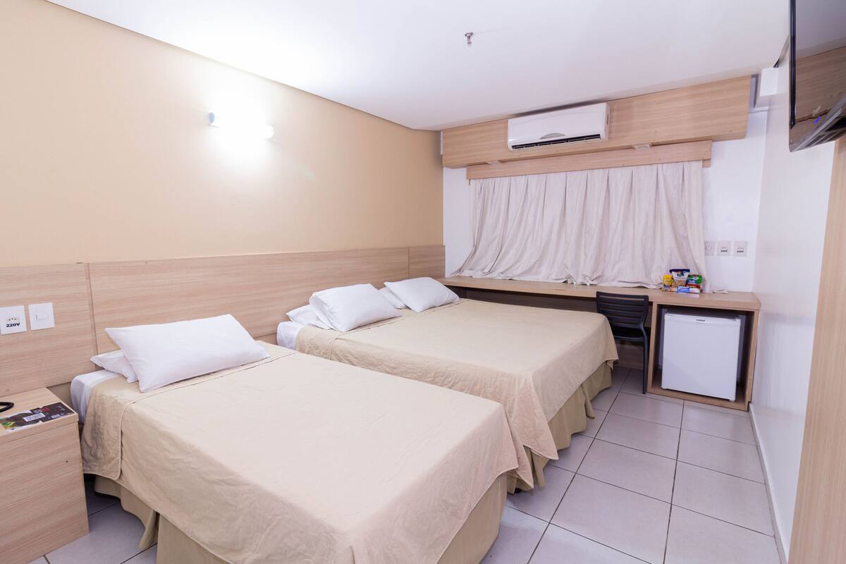A hotel room with two beds, a desk, a chair, a small refrigerator, and an air conditioning unit.