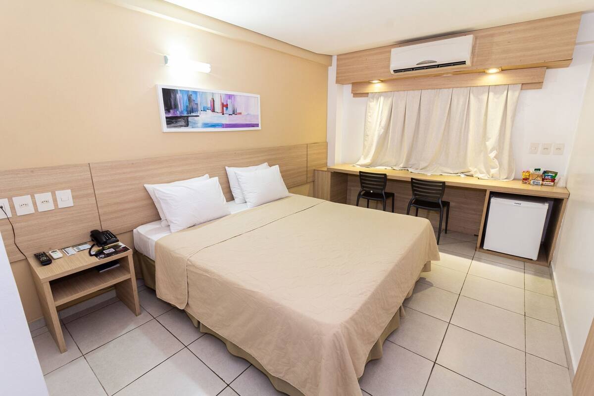 A hotel room with a bed, bedside table, desk, chairs, and a small refrigerator.
