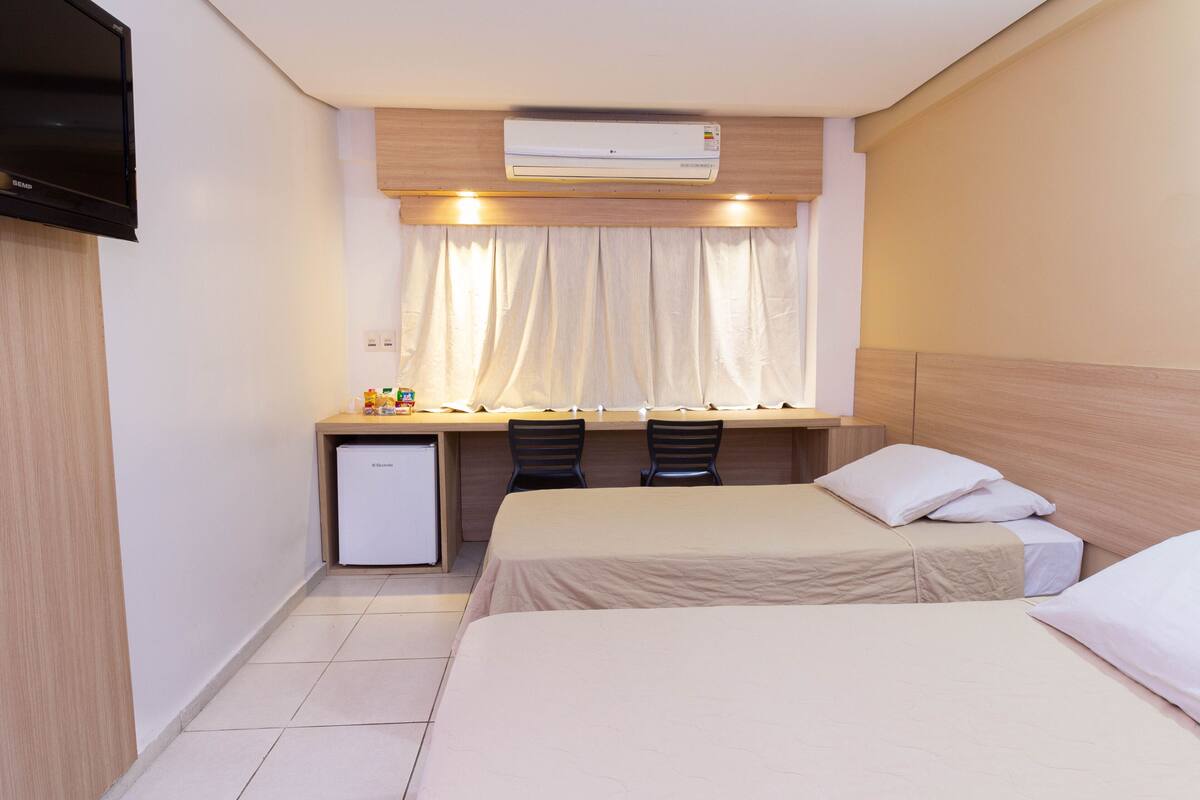 A compact hotel room with two single beds, a small desk with two chairs, a television, and an air conditioning unit.