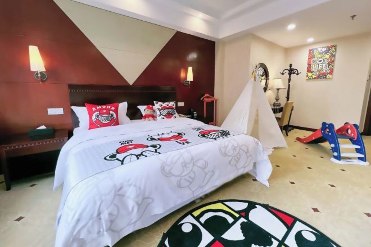 A hotel room with a bed featuring a white bedspread with cartoon characters, a wooden headboard, two wall-mounted lamps, a small table with a lamp, a chair, and a colorful rug.
