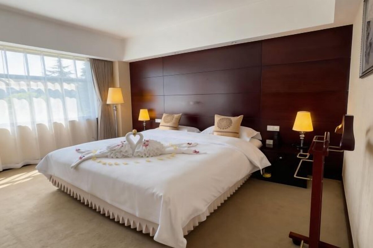 Une chambre d’hôtel avec un grand lit, deux lampes de chevet, une tête de lit sombre et une fenêtre avec des rideaux.