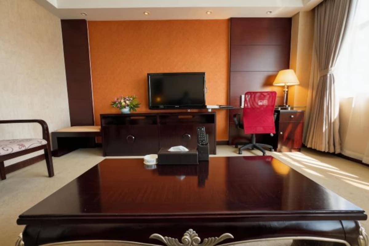 A hotel room with a wooden desk, a television, a red chair, a lamp, and a small plant.