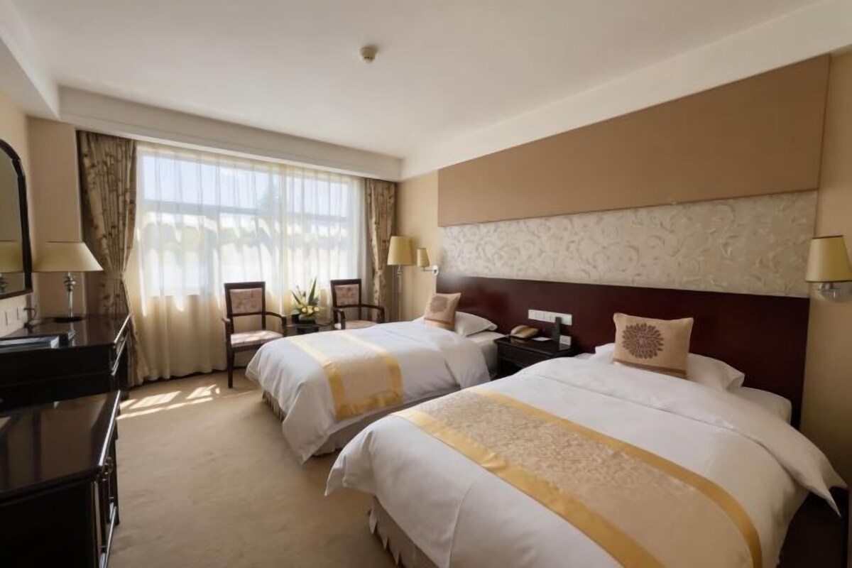 A hotel room with two beds, a desk, a chair, a nightstand, and a large window with curtains.