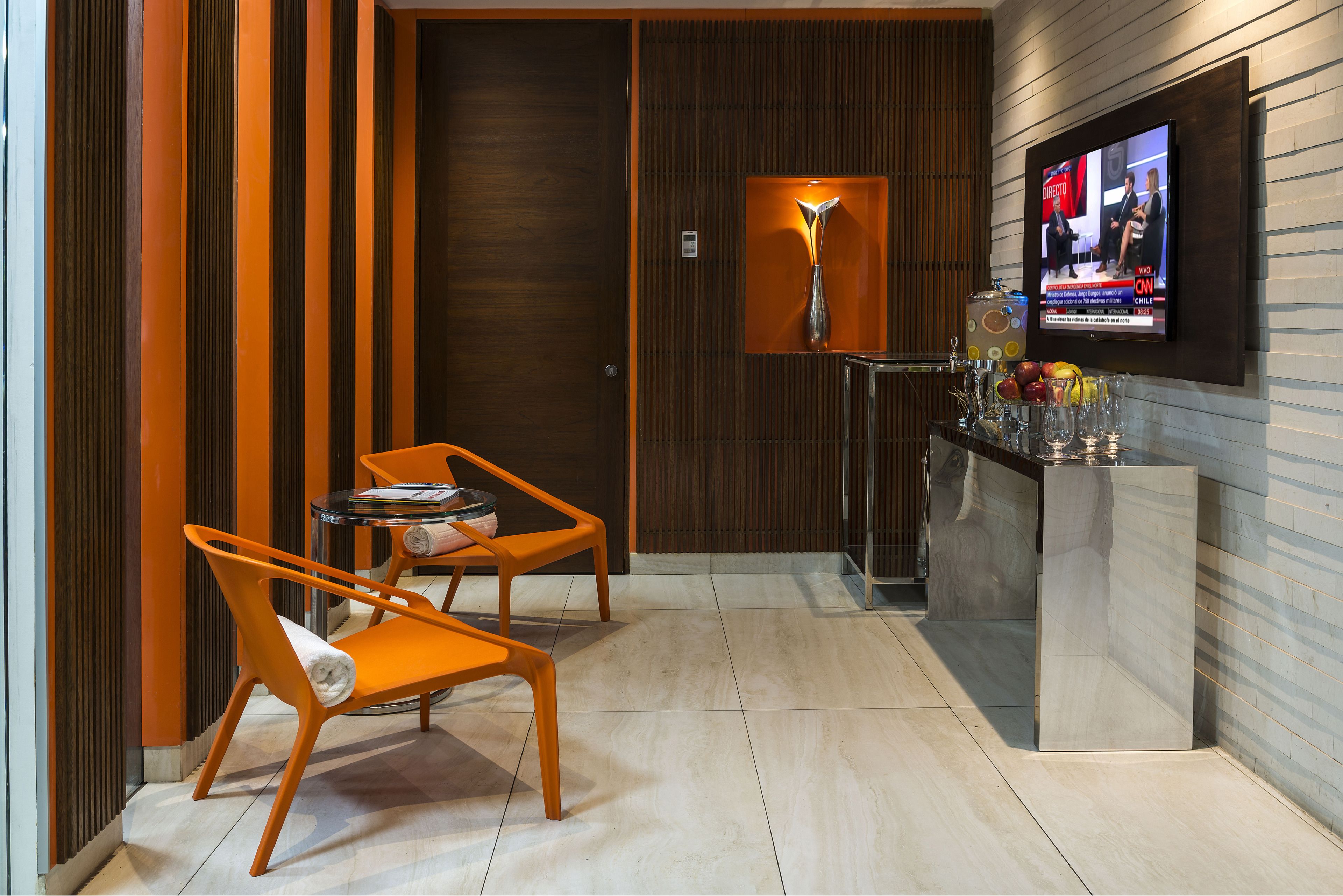 Quarto de hotel moderno com parede de painel de madeira, TV fixada na parede, mesa redonda pequena com uma cadeira e um armário de vidro com frutas.