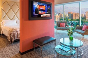 Habitación de hotel con cama, televisión montada en la pared, zona de estar con sofá y sillones, y vistas a un paisaje urbano a través de amplias ventanas.