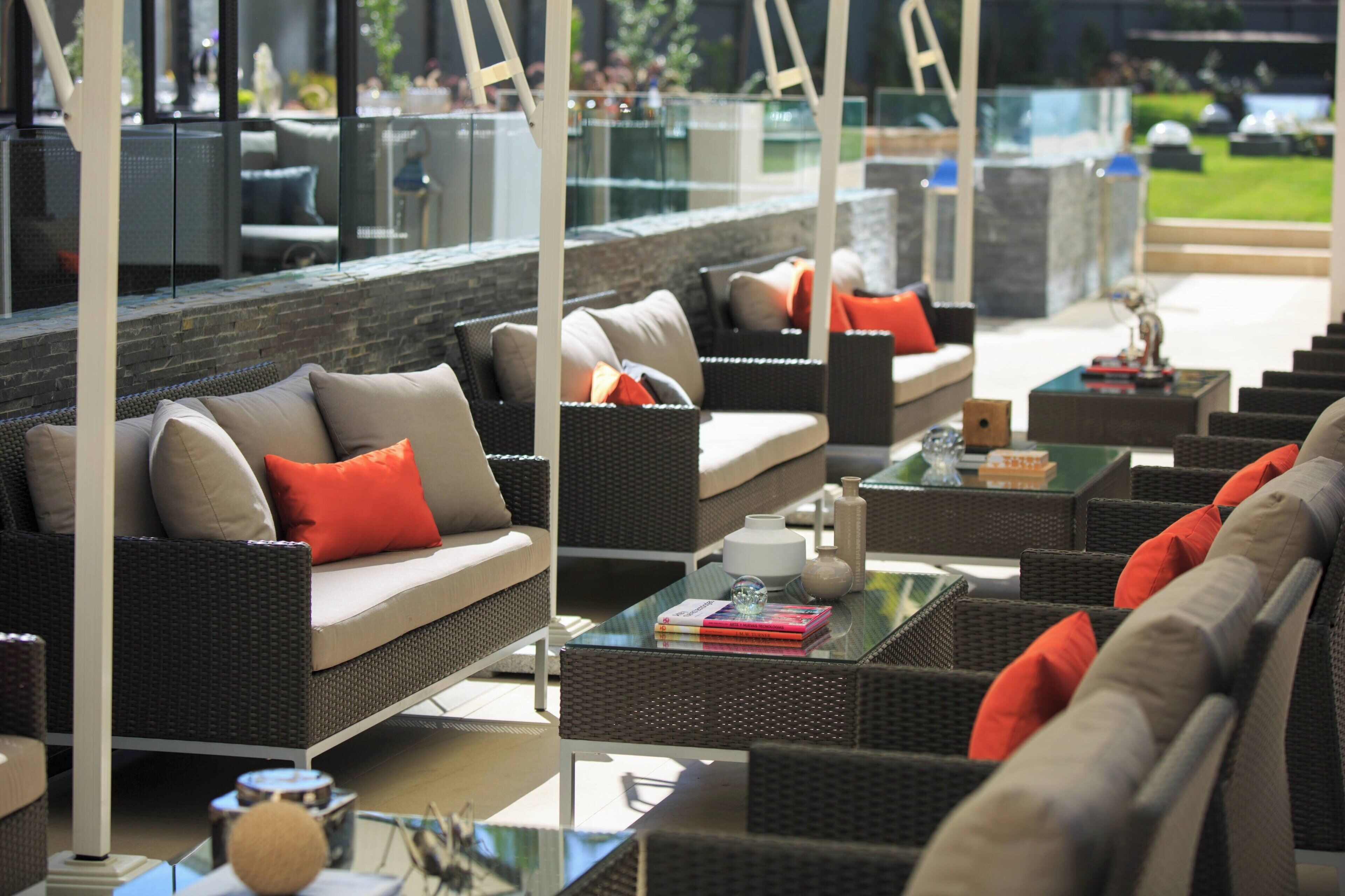 A modern outdoor seating area with wicker furniture and glass tables, featuring a stone wall and a view of a green landscape.