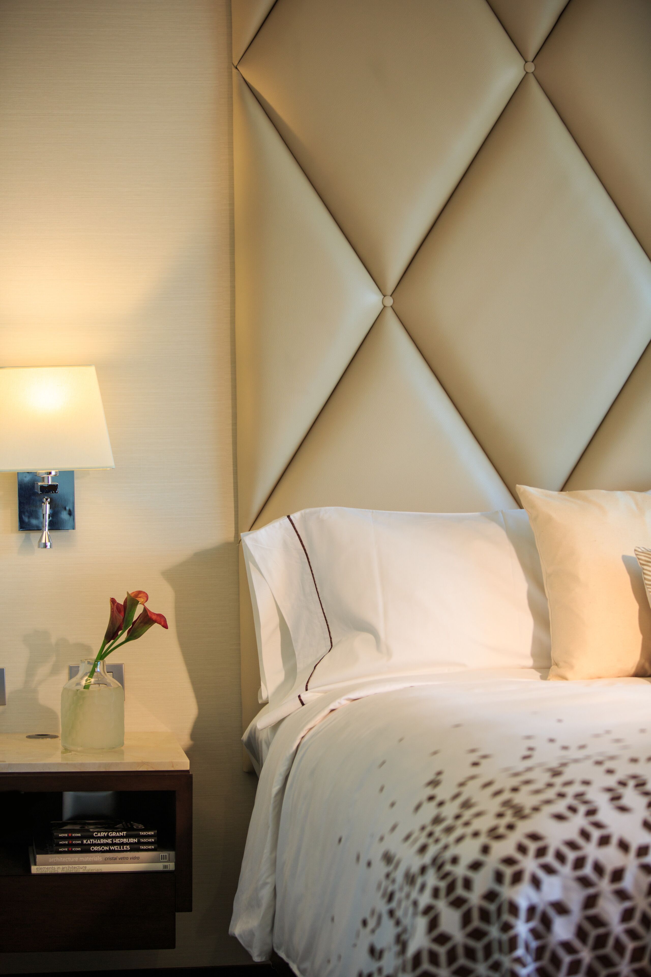 Habitación de hotel con un cabecero capitonado, una mesita de noche con lámpara, un jarrón con flores y una cama con colcha estampada.