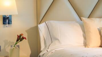 Habitación de hotel con un cabecero capitonado, una mesita de noche con lámpara, un jarrón con flores y una cama con colcha estampada.