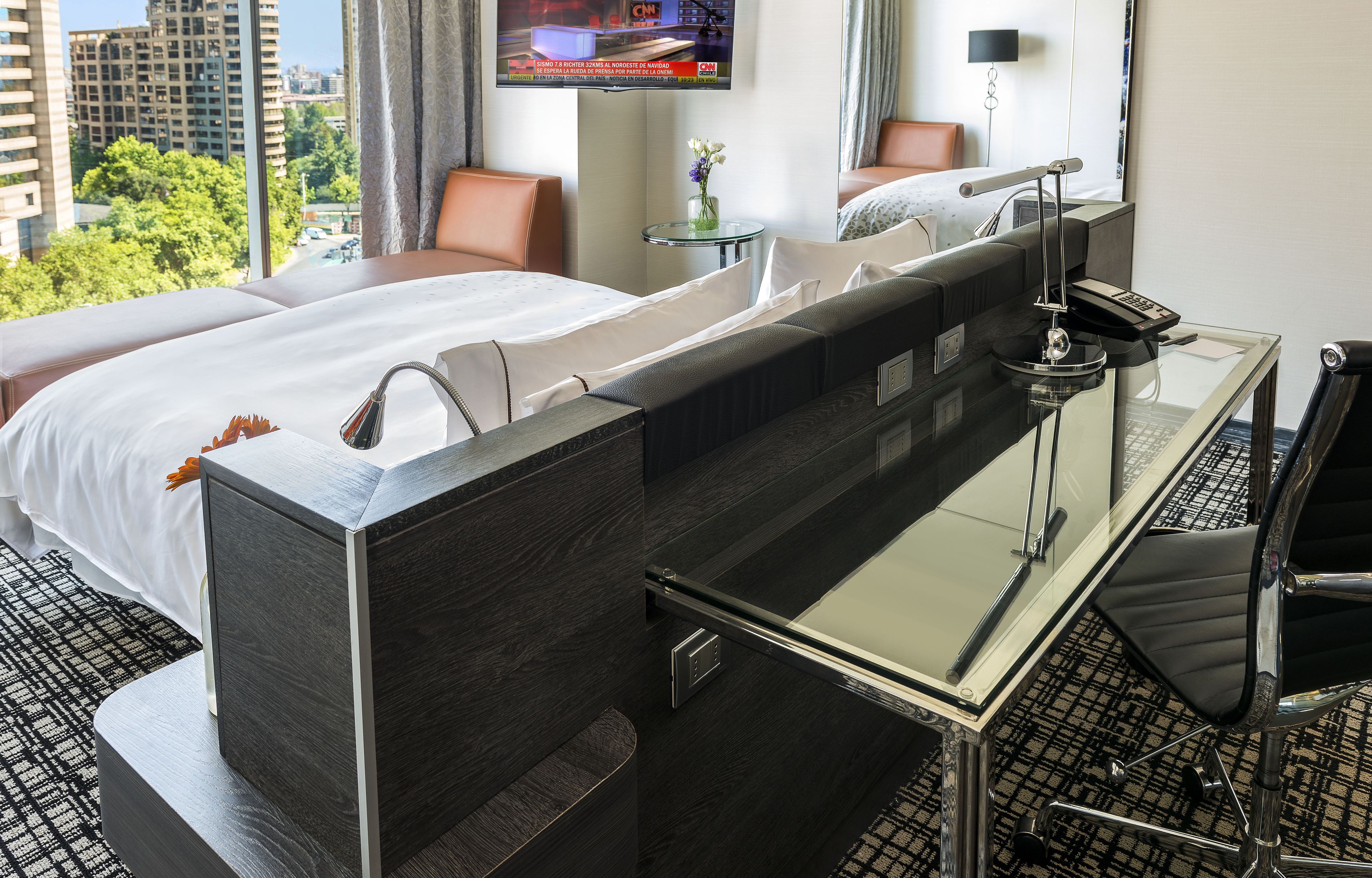 A hotel room with a large bed, a desk with a computer, a chair, a TV mounted on the wall, and a view of the cityscape.