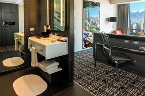 A modern hotel bathroom with a large mirror, a sink, and a toilet. There is a chair and a desk with a view of the city.