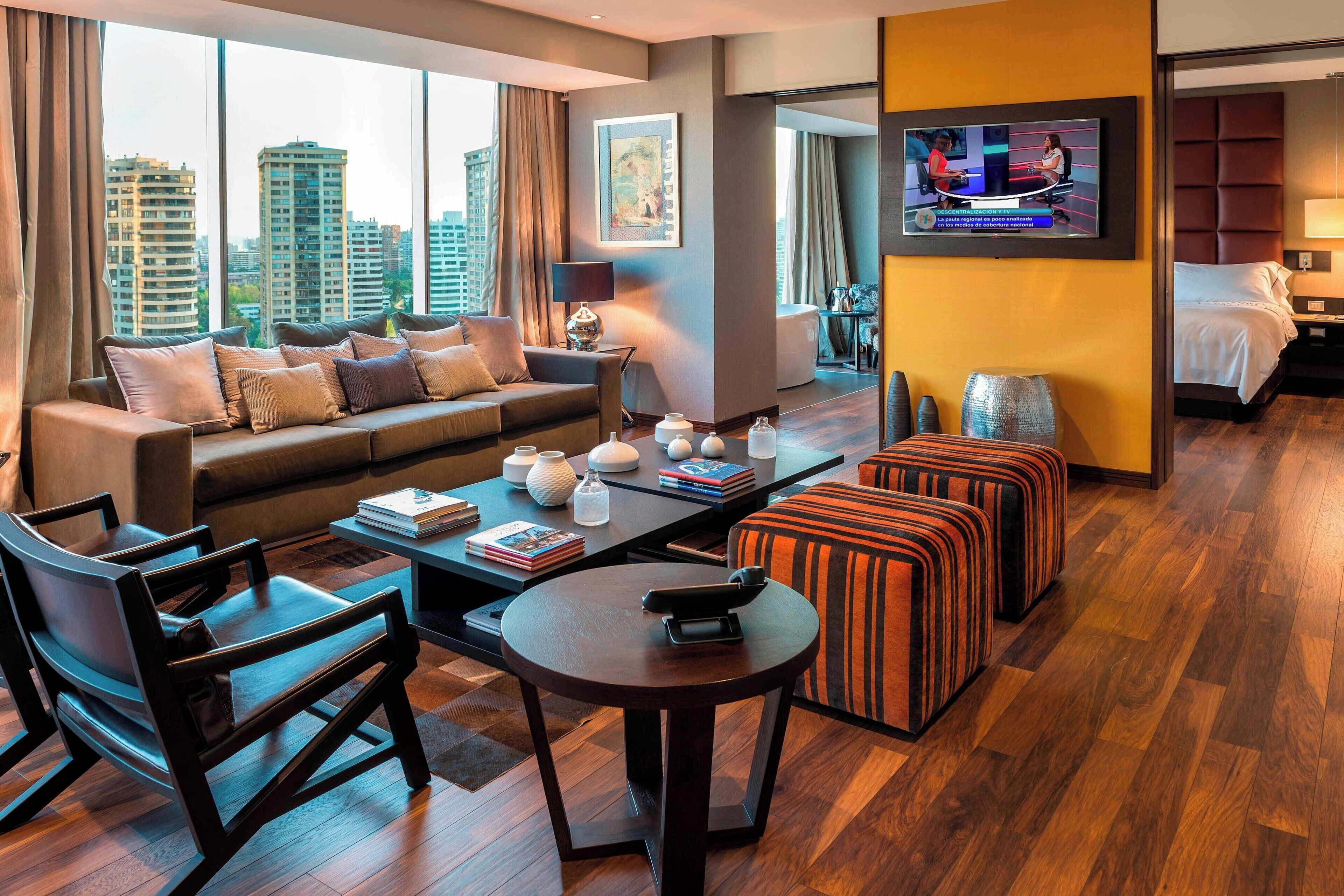 A modern hotel room with a large window, a sofa, a coffee table, a TV, and a bed.