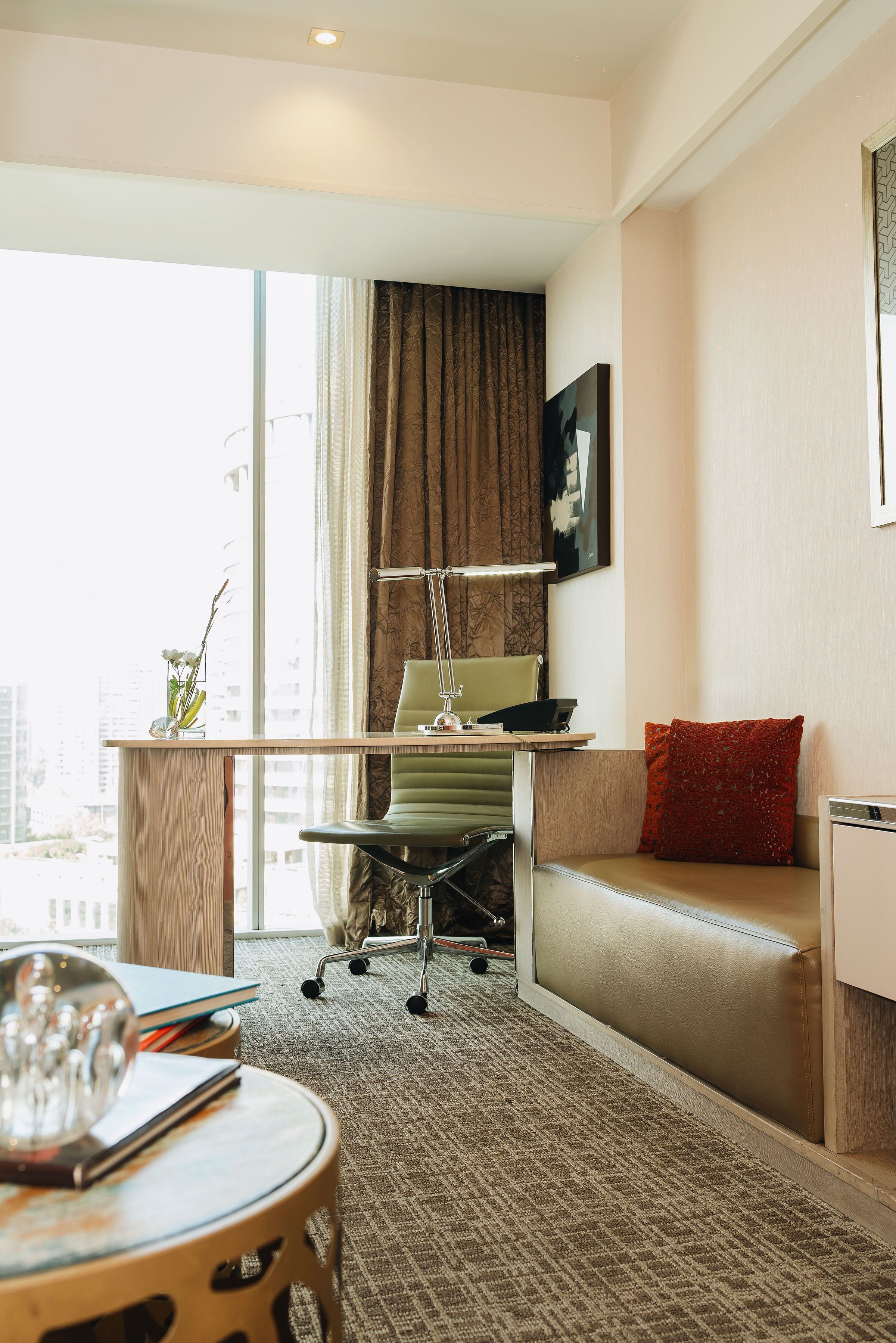 Una habitación de hotel moderna con un ventanal grande, un escritorio con una computadora portátil, una silla, un sofá y una pequeña mesa con un adorno.