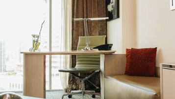 Una habitación de hotel moderna con un ventanal grande, un escritorio con una computadora portátil, una silla, un sofá y una pequeña mesa con un adorno.
