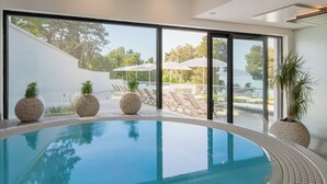 Une piscine intérieure moderne aux parois en verre transparent, entourée de chaises longues et de plantes en pot.