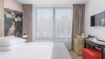 Et hotelværelse med en stor seng, et skrivebord med stol, et fjernsyn og et vindue med let gardin.