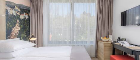 A hotel room with a large bed, a desk with a chair, a television, and a window with sheer curtains.