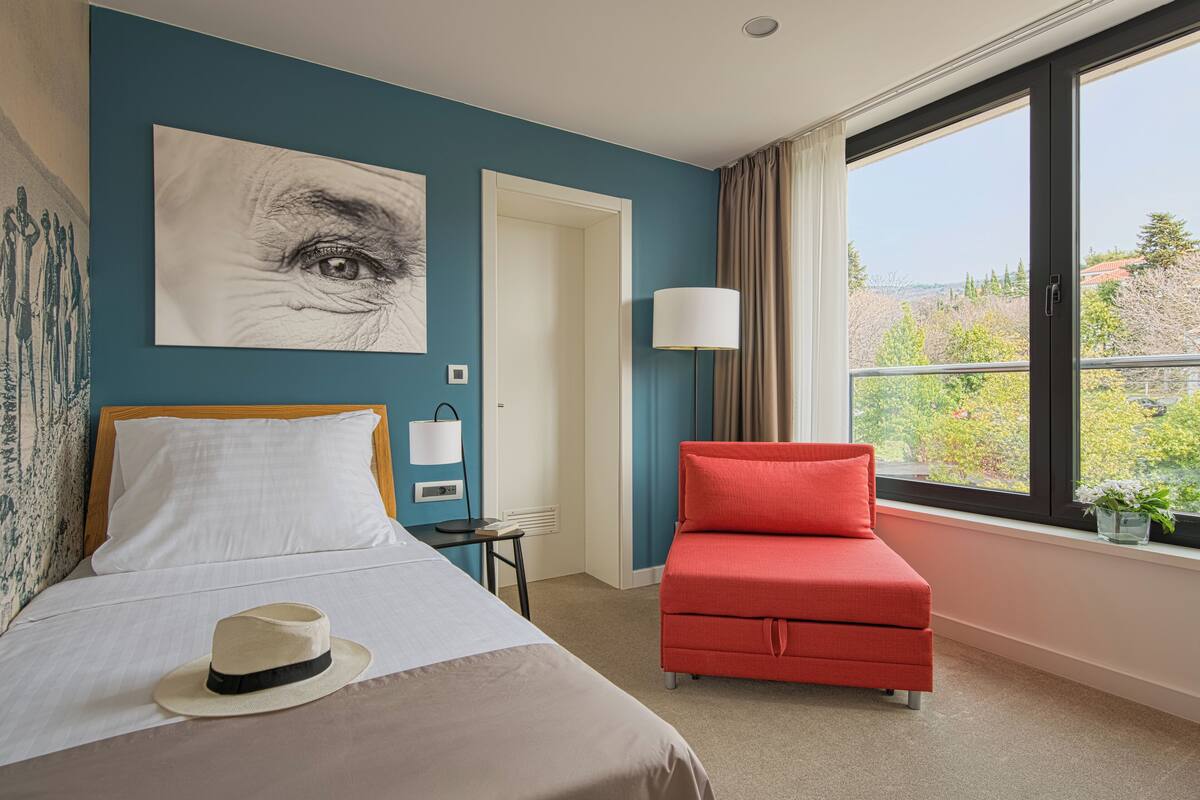 Une chambre d’hôtel avec un lit, un fauteuil rouge, une fenêtre donnant sur un paysage verdoyant et un tableau accroché au mur.