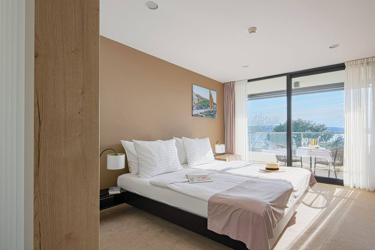 Une chambre d’hôtel moderne avec un grand lit, une table de chevet avec une lampe, une vue sur le balcon et un tableau au mur.