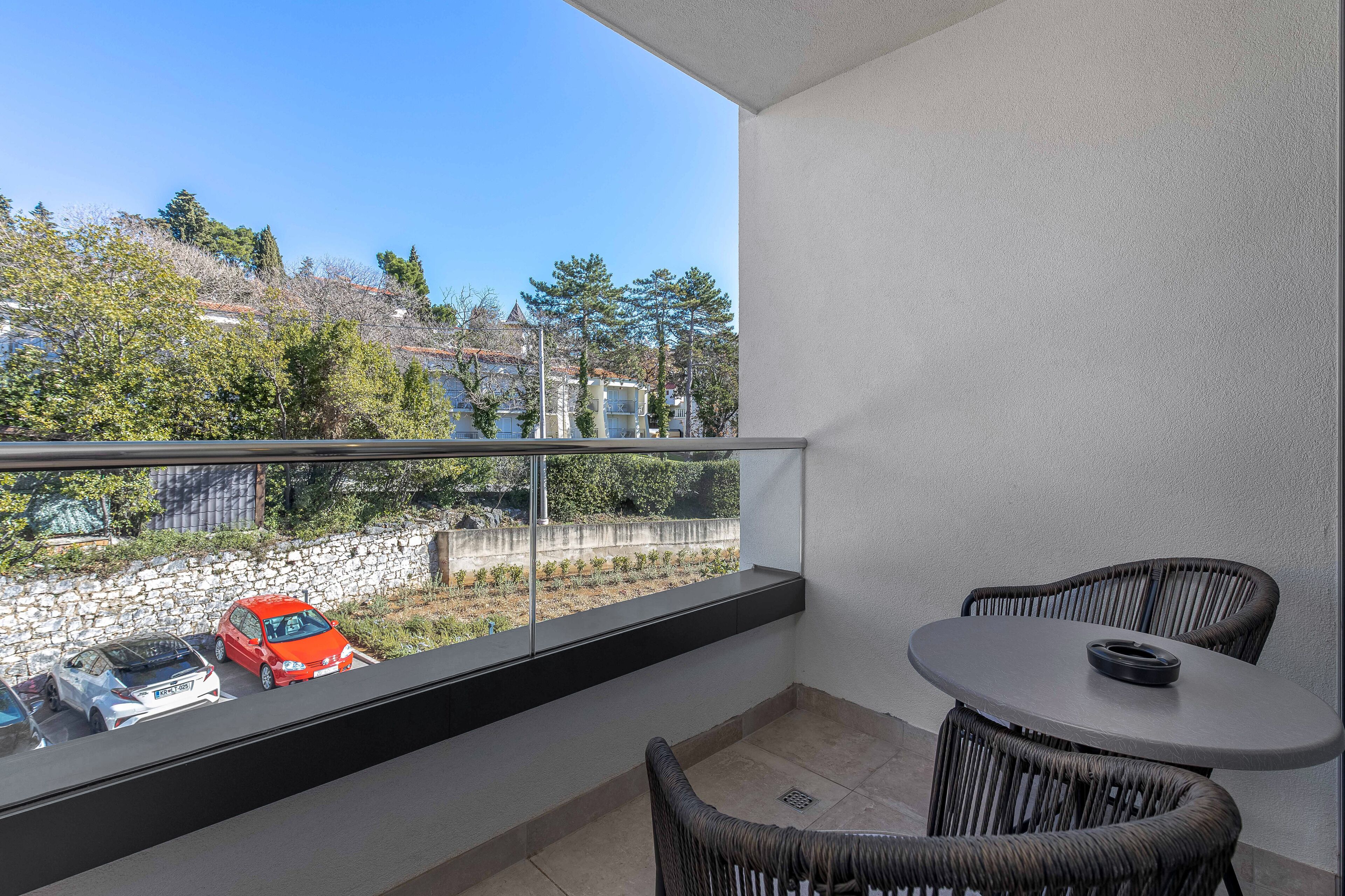 A balcony with a table and chairs, a stone wall, and a view of parked cars and trees.