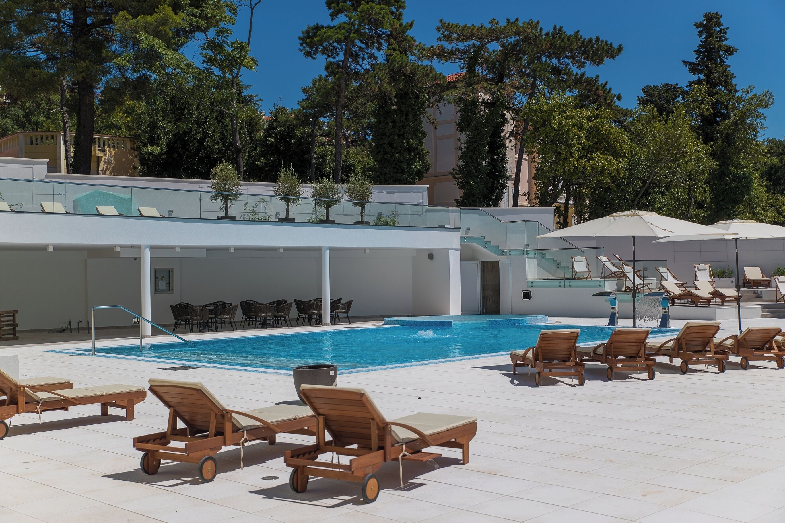 A pool area with wooden lounge chairs and umbrellas, surrounded by trees and a covered seating area.