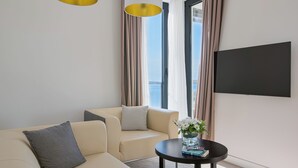 Une chambre d’hôtel moderne avec un canapé beige, une table basse ronde ornée d’un vase de fleurs, un téléviseur à écran plat fixé au mur et une grande fenêtre avec des rideaux.