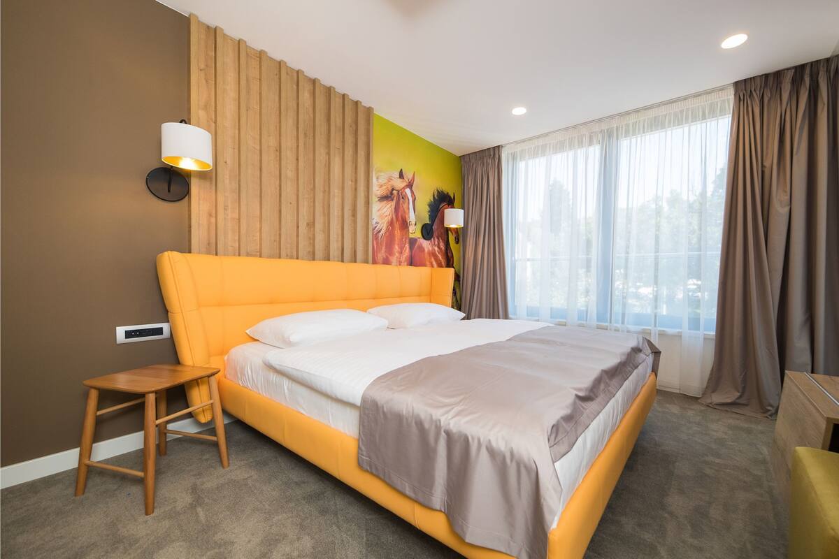 Une chambre d’hôtel avec un grand lit, une petite table en bois, une lampe fixée au mur et une grande fenêtre avec des rideaux.